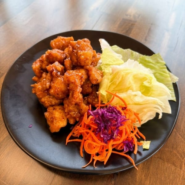 Crispy orange chicken with fresh lettuce wraps, shredded carrots, and purple cabbage on a black plate.