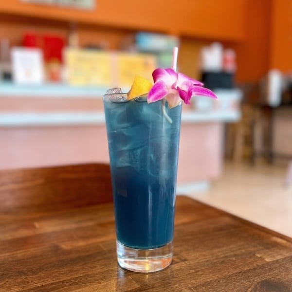 Blue cocktail with ice, a lemon wedge, and a pink orchid garnish on a wooden table.