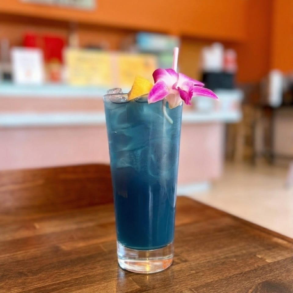 Blue cocktail with ice, a lemon wedge, and a pink orchid garnish on a wooden table.